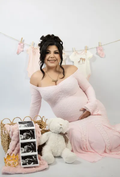 Smiling pregnant woman in pink dress sitting with ultrasound photos teddy bear and baby clothes.
