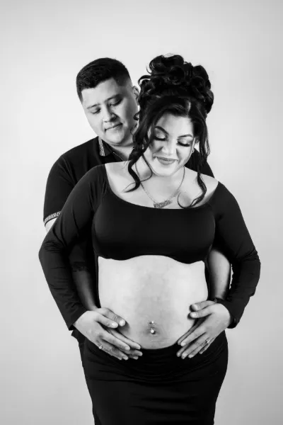 Black and white portrait of couple cradling pregnant belly together against light background.