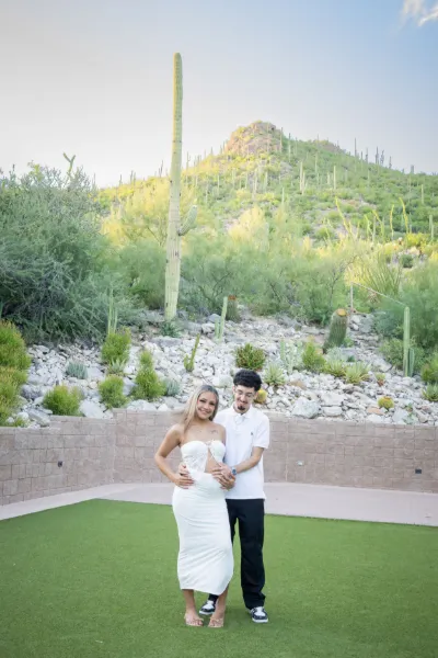 Couple posing together on green lawn with saguaro cactus hillside backdrop during maternity photo session.