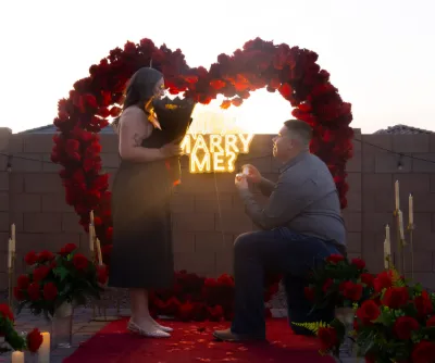 Man proposes marriage on one knee surrounded by red roses and heart-shaped lights at sunset.
