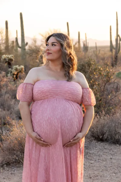 Pregnant woman in pink off-shoulder lace dress cradling baby bump at golden hour in desert with saguaro cacti.