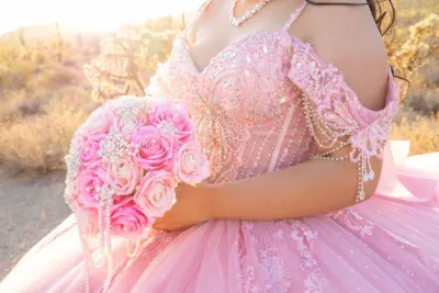 Young woman in pink beaded quinceañera ball gown holds matching pink rose bouquet in golden sunlight.