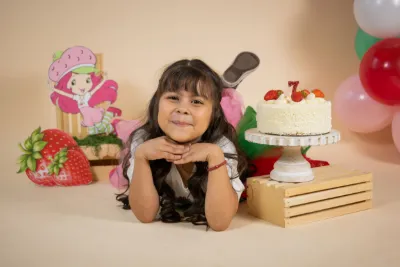 Young girl poses with Strawberry Shortcake themed birthday cake and decorations celebrating her 7th birthday.