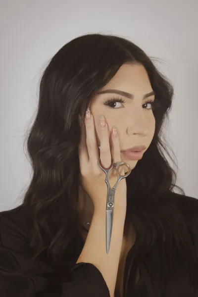 Brunette woman with long wavy hair holding scissors near her face against a light gray background.