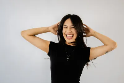 Laughing young woman in black t-shirt with hands in hair against white background.