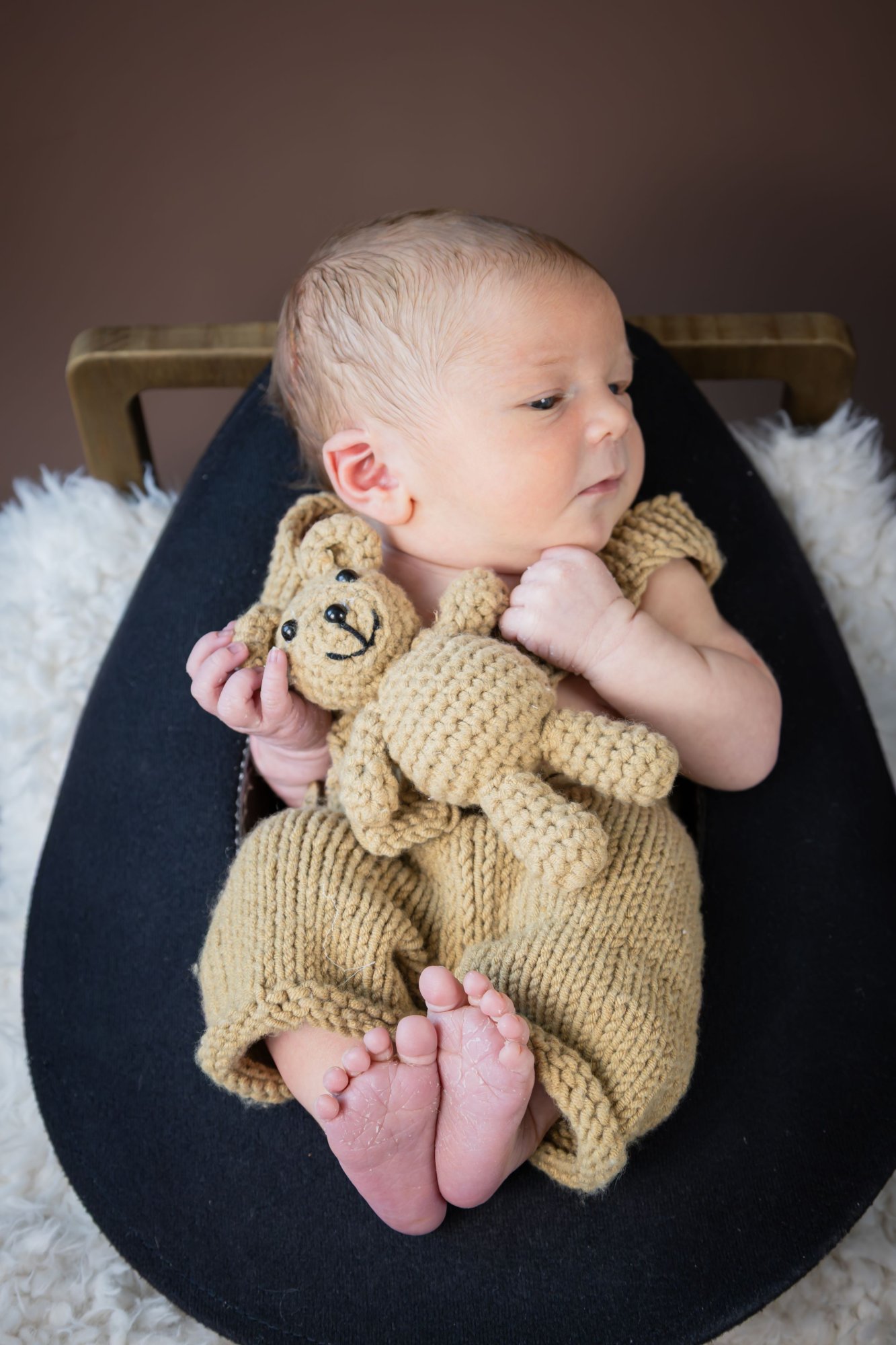 Newborn baby in knit tan romper lying on dark cushion holding a small crocheted teddy bear.