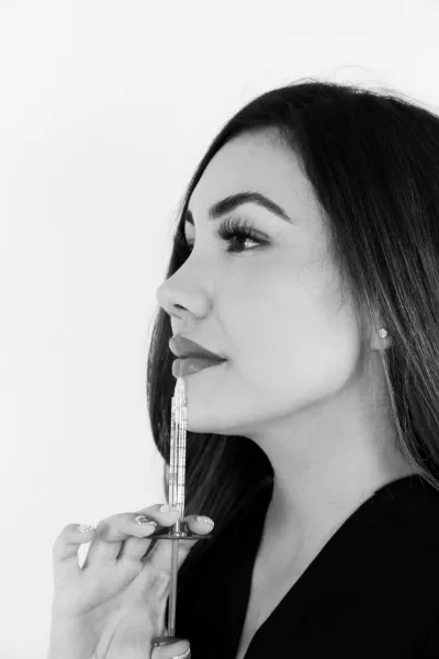 Black and white profile portrait of a young woman holding a syringe near her lips against white background.