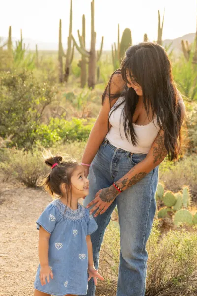 A tattooed mother smiles down at her young daughter in a blue dress among saguaro cacti in the Arizona desert.