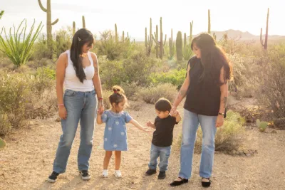 Two women hold hands with two young children walking together through a sunny saguaro cactus desert landscape.