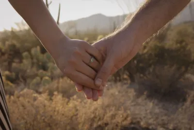A close-up of a couple holding hands in the desert, showcasing an engagement ring among desert shrubs.