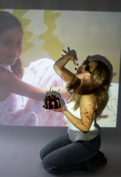 Woman kneeling eating cake in front of projected childhood photo in creative birthday shoot.