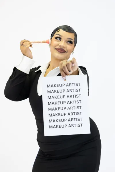 A makeup artist in a black suit holds a sign reading MAKEUP ARTIST repeatedly while pointing a brush at her temple.