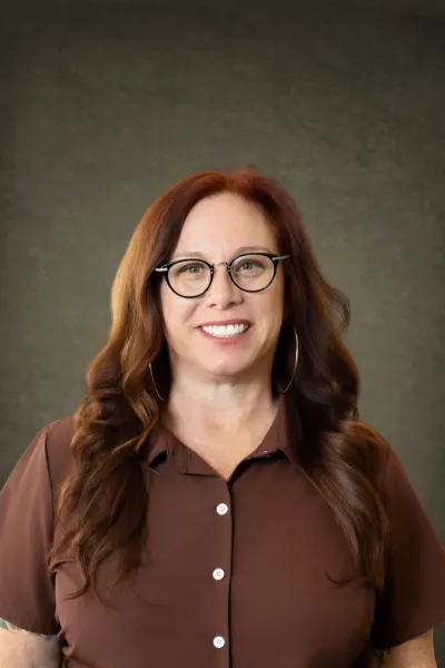Smiling woman with red hair and glasses wearing a brown shirt poses for a professional headshot photo.