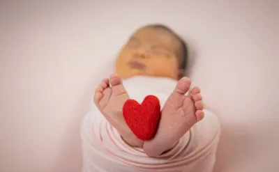 Newborn baby feet holding a small red felt heart while swaddled in a pink wrap blanket.