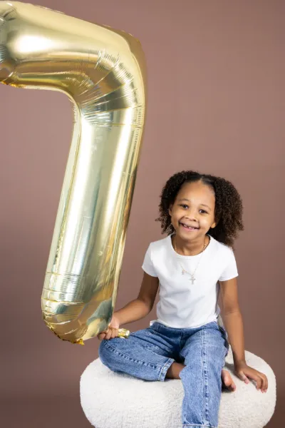 Happy young Black girl sitting on white pouf holding gold number 7 balloon on brown background.