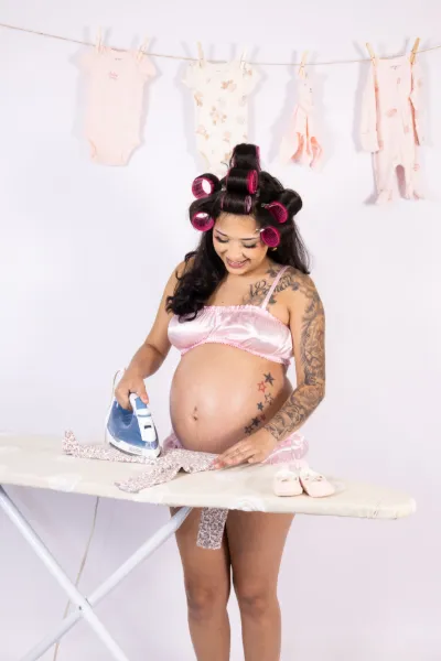 Tattooed pregnant woman in pink lingerie with hair curlers ironing baby clothes with laundry hanging in background.