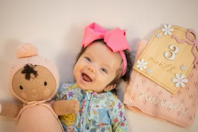 A happy 3-month-old baby girl in a floral outfit smiles beside a stuffed doll and milestone card.