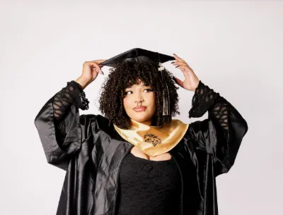 A young woman in black graduation cap and gown with gold hood posing against a white background.