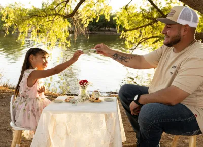 A young girl in a pink dress shares a sweet tea party moment with her father by a peaceful riverside.