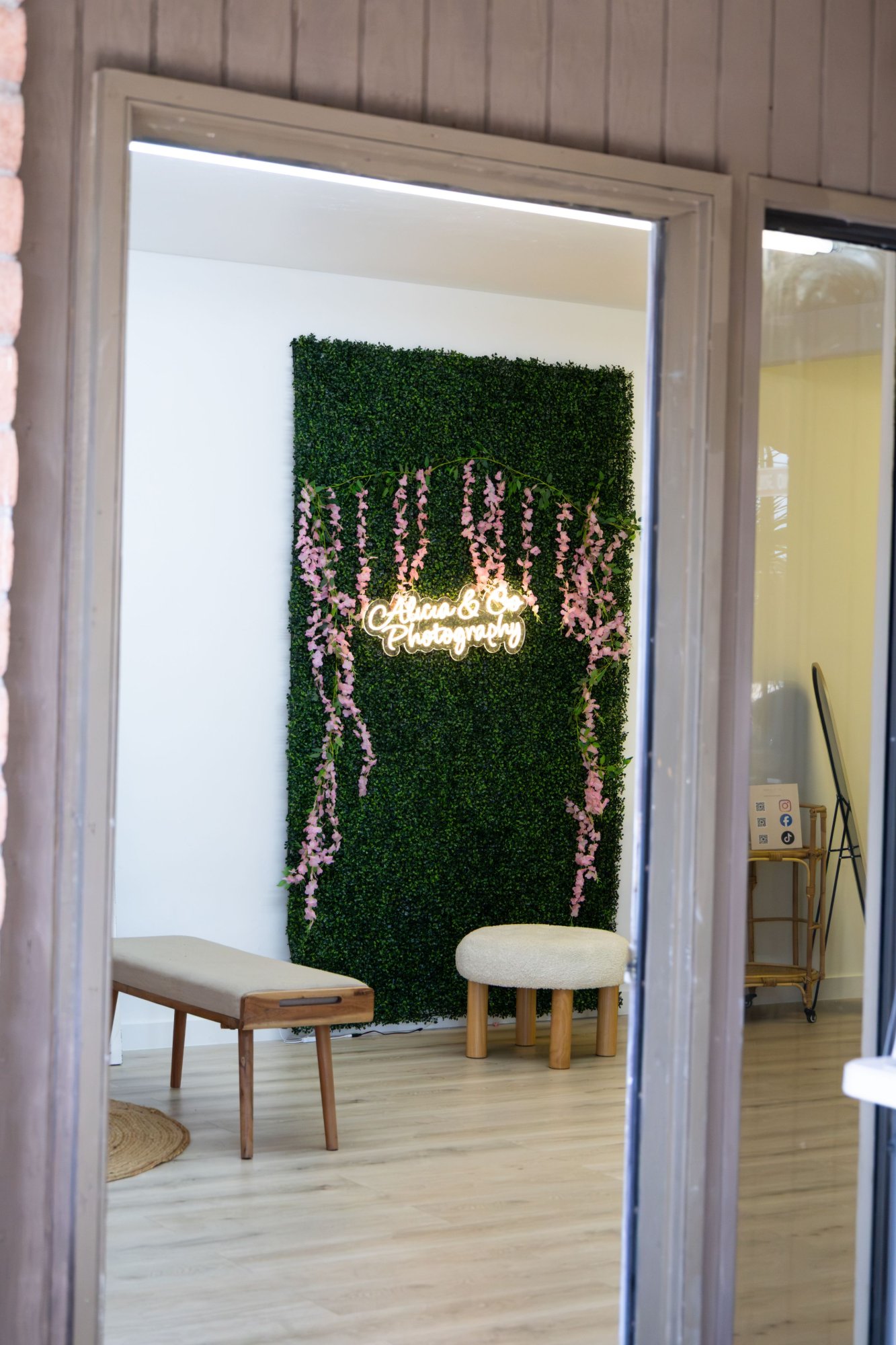 The Alicia & Co. studio in Tucson: the neon script sign on a green boxwood wall with pink wisteria, a bench and a stool.